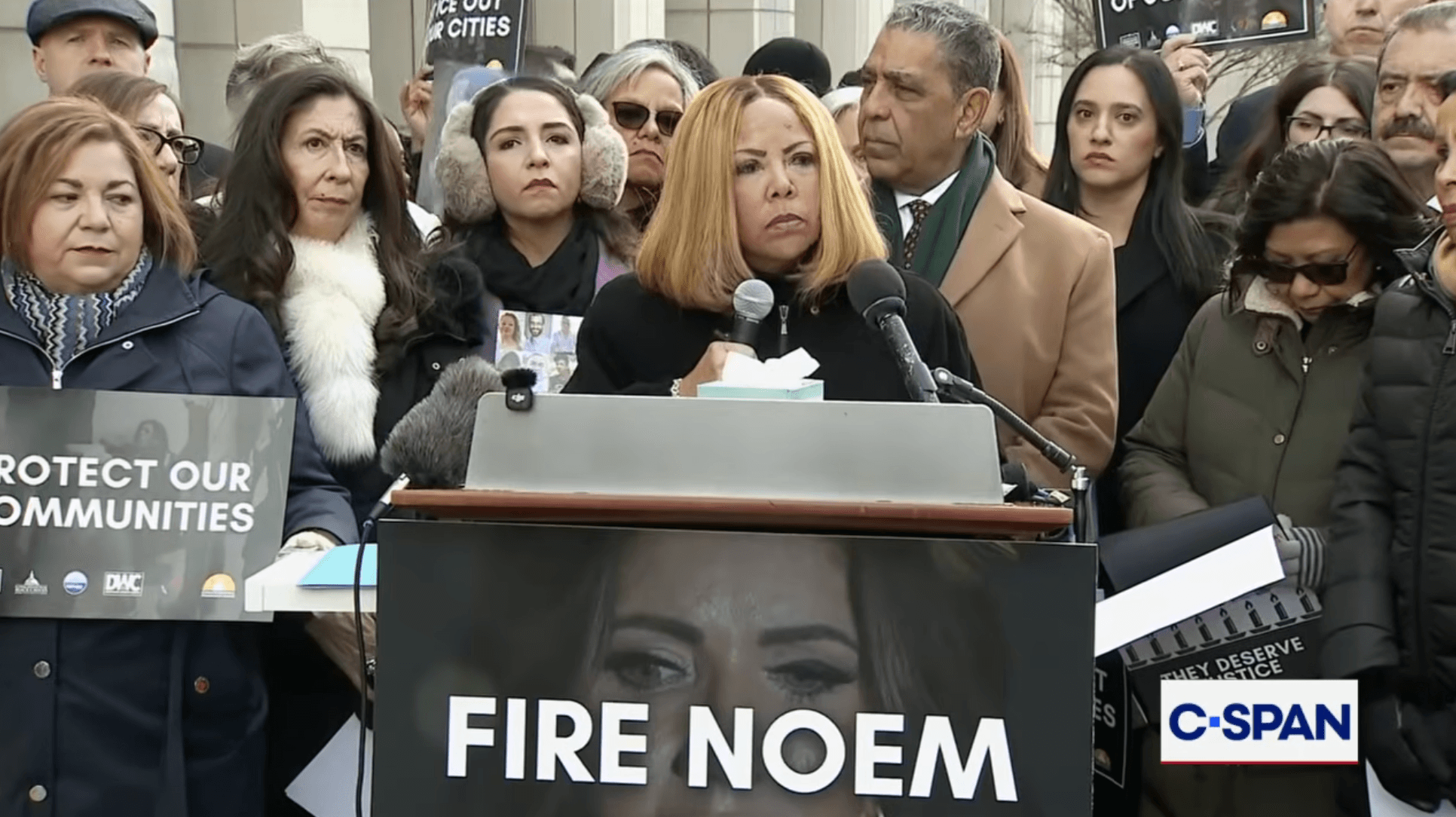Image of Rep. Lucy McBath standing outdoors at a podium holding a microphone. On the podium is a sign that reads "Fire Noem."