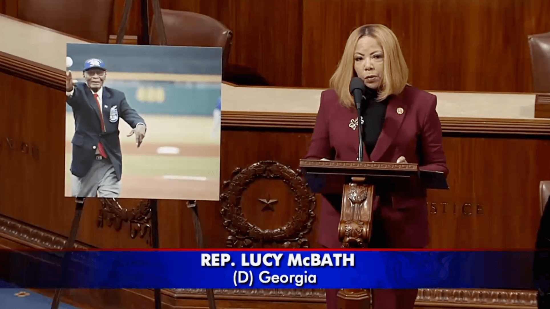 Photo of Rep. McBath speaking from the floor of the U.S. House of Representatives. She is standing next to a picture of Dr. Hillard Pouncy, Jr. throwing out the first pitch at a baseball game.