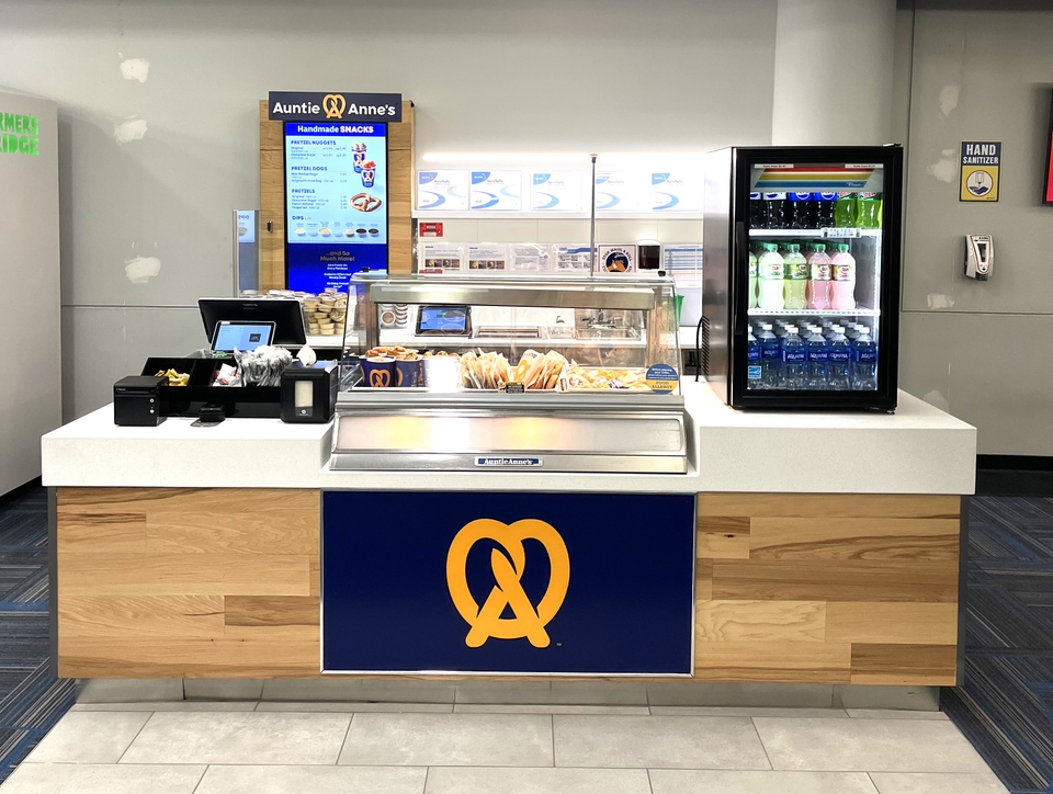 Auntie Anne's pretzel stand in an indoor setting, featuring a display of pretzels, a digital menu, and a refrigerator with beverages.