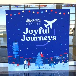 Promotional display at Reagan National Airport featuring the text 'Joyful Journeys' with a colorful illustration of a winter holiday scene at an airport, including travelers and an air traffic control tower.