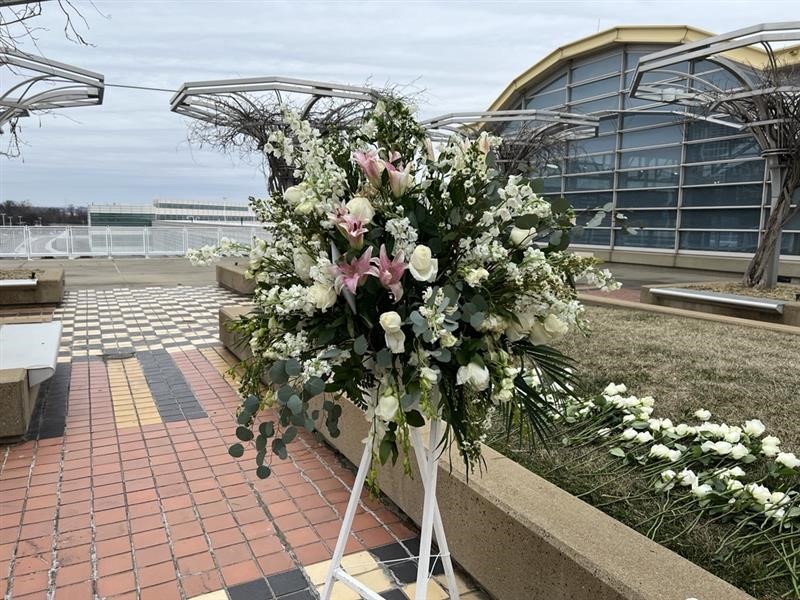 A large floral arrangement stands on a stand in a paved plaza with curved benches and DCA Airport architectural structures in the background.