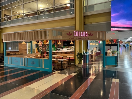 Newly opened vibrant café named "Colada Shop" with teal and wood accents, located in a spacious indoor setting at Reagan National Airport. The café features a bar, seating area, and decorative plants, creating an inviting atmosphere.