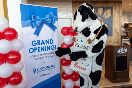 A person in a cow costume is standing next to a "Grand Opening!" sign at a Chick-fil-A event in Reagan National Airport. The sign is decorated with a large blue ribbon, and there are red and white balloons nearby.