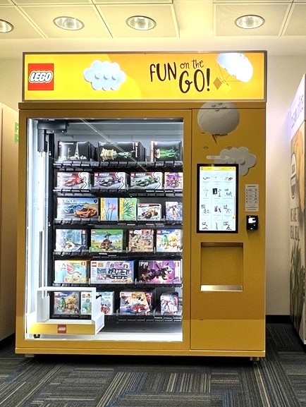 A brightly lit LEGO vending machine is labeled "FUN on the GO!" and filled with various LEGO sets at Dulles International Airport. There's a digital screen on the right side for selections. The machine is housed in a clean, well-lit space with a carpeted floor.