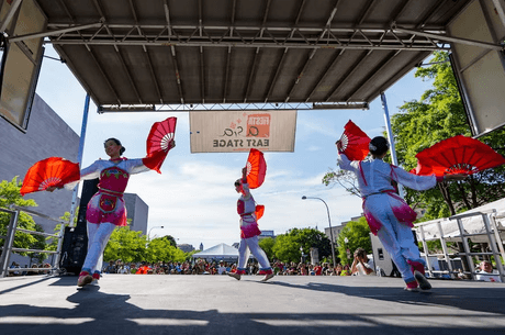 A lively outdoor performance at a festival, with individuals on stage wearing vibrant costumes and holding red fans. A sign above reads "Fiesta Asia East Stage." A large audience watches from the background, and the scene is framed by trees and buildings.