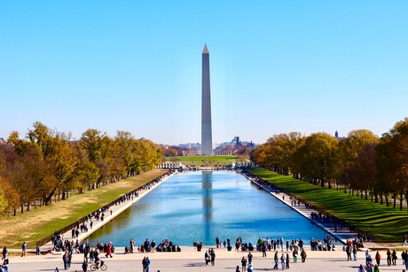 The image shows the Washington Monument in the center, with a reflecting pool leading towards it. The scene includes numerous visitors walking along the pathways flanking the water. The sky is clear, and trees line both sides of the pool.