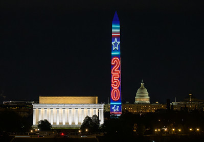 The image shows the Washington Monument illuminated with colorful lights displaying "250" and stars. The Lincoln Memorial is in the foreground, lit up against the night sky. The U.S. Capitol building is visible in the background.