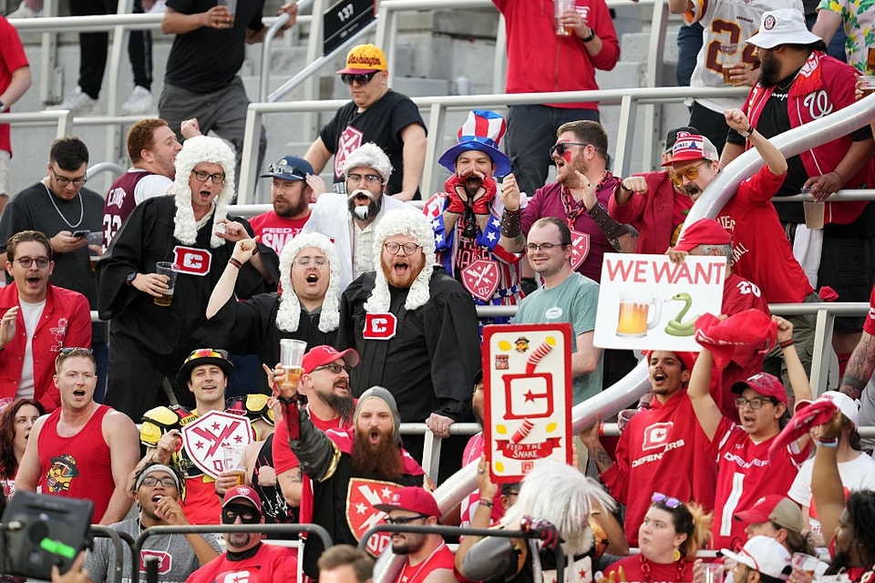 A lively crowd of DC Defenders football fans, dressed in various costumes, cheer in stadium seats. Some are wearing wigs resembling judges’ attire. Several hold signs.