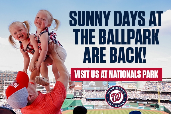 A person holds up two smiling children at a baseball game. The sign reads, "Sunny days at the ballpark are back! Visit us at Nationals Park." The Washington Nationals logo is visible.