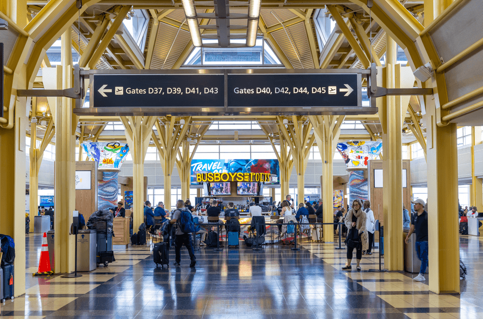 A bustling airport terminal with travelers and luggage. A sign for "Busboys and Poets" is visible in the background amidst yellow architectural beams and large windows at Reagan National Airport.