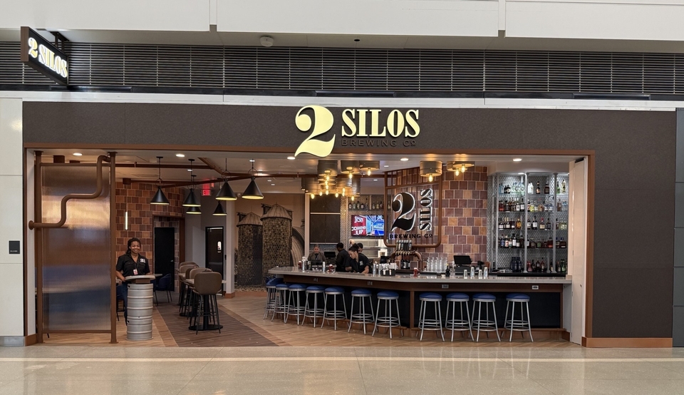 The image shows the "2 Silos Brewing Co." bar in Dulles International Airport terminal. It features a wide counter with several bar stools, a variety of bottles on shelves, and cushioned seating in the background. A person is standing behind the bar counter.