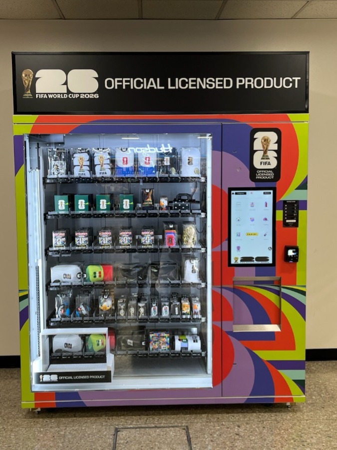 A colorful vending machine branded with FIFA World Cup 2026 logos, displaying various official licensed products, including small merchandise and collectibles. An interactive screen is positioned on the right side.