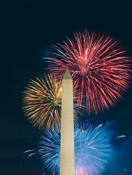 The Washington Monument at night with colorful fireworks in red, yellow, and blue lighting up the sky behind it.
