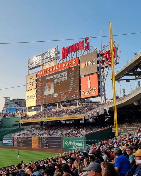 A baseball game at Nationals Park with the scoreboard prominently displaying player information. The stadium is filled with spectators. Logos for PNC and Coca-Cola are visible, and advertisements from Geico and National Car Rental are displayed around the field.