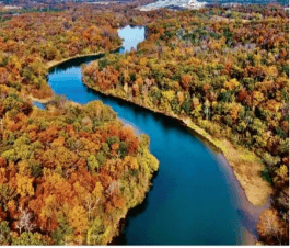 Aerial view of a river winding through a forest with vibrant autumn foliage, showcasing shades of red, orange, and yellow. The sky is clear and blue, enhancing the natural beauty of the scene.