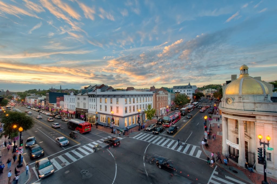 A bustling street scene in Georgetown, Washington D.C., featuring historic buildings and a prominent domed structure on the right. The sky is partly cloudy with a vibrant sunset.