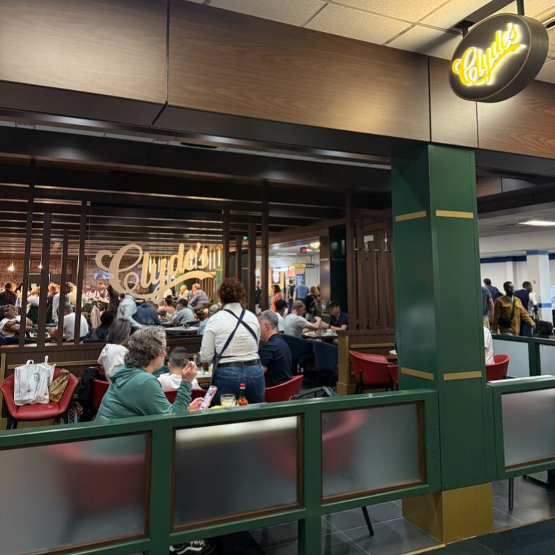 A busy dining area at Clyde's restaurant at Dulles International Airport, with many people seated at red chairs around tables. A sign with the Clyde's logo is visible above. There are wooden dividers and a ceiling with recessed lighting.
