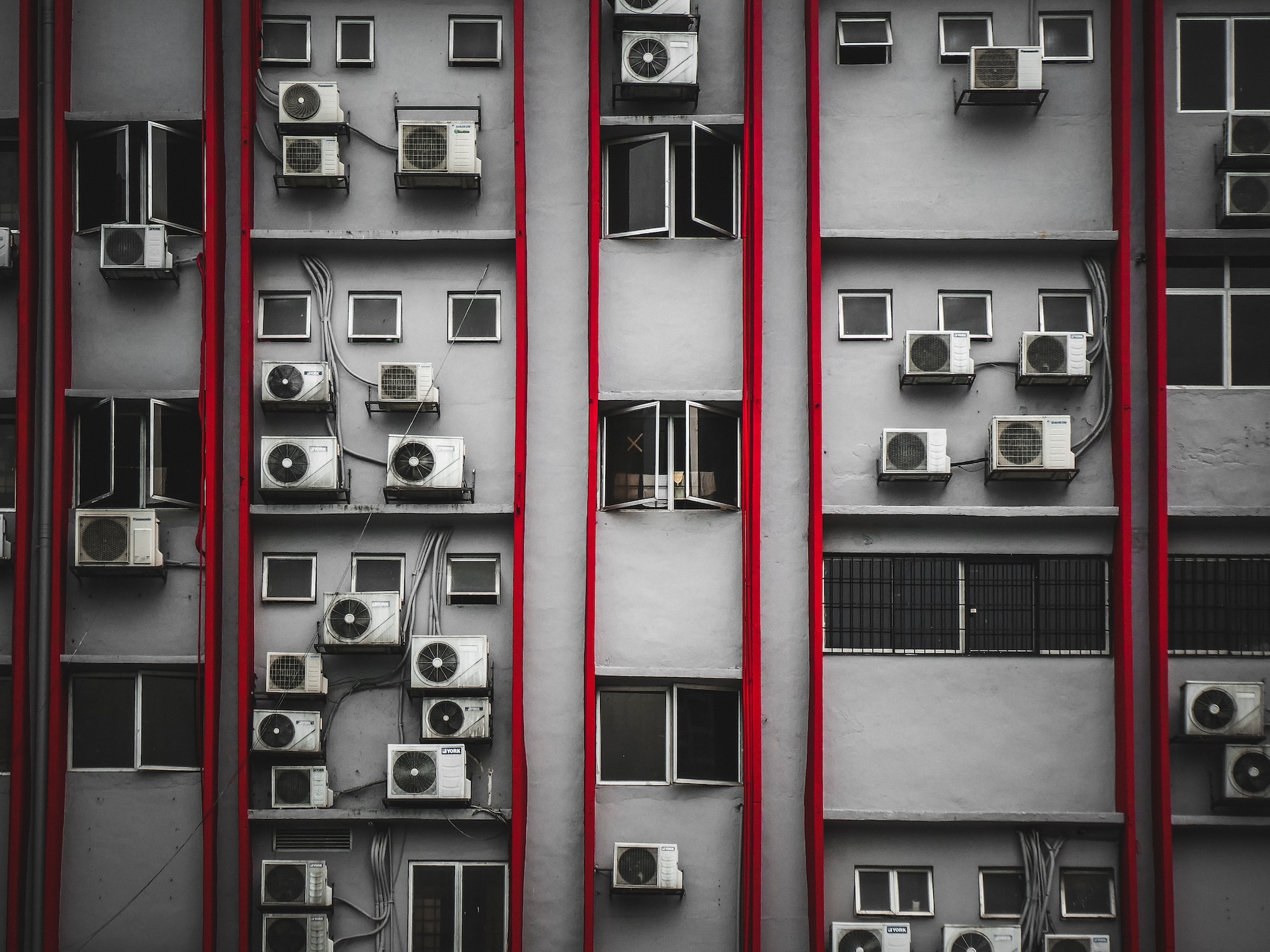 Gri bir apartman duvarında kırmızı su boruları ve her katta birkaç tane klima görülüyor.