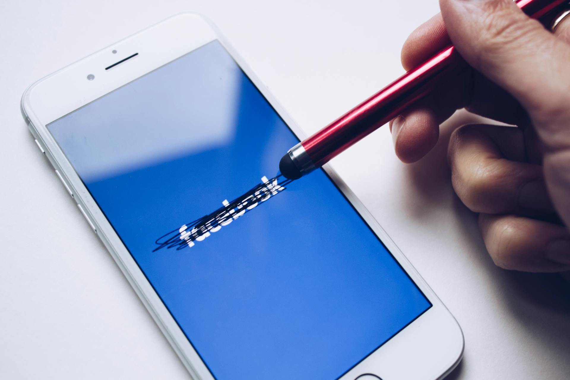 Beyaz bir iPhone 6'nın ekranında Facebook logosu var. Birisi elindeki stylus ile logonun üzerini karalıyor.