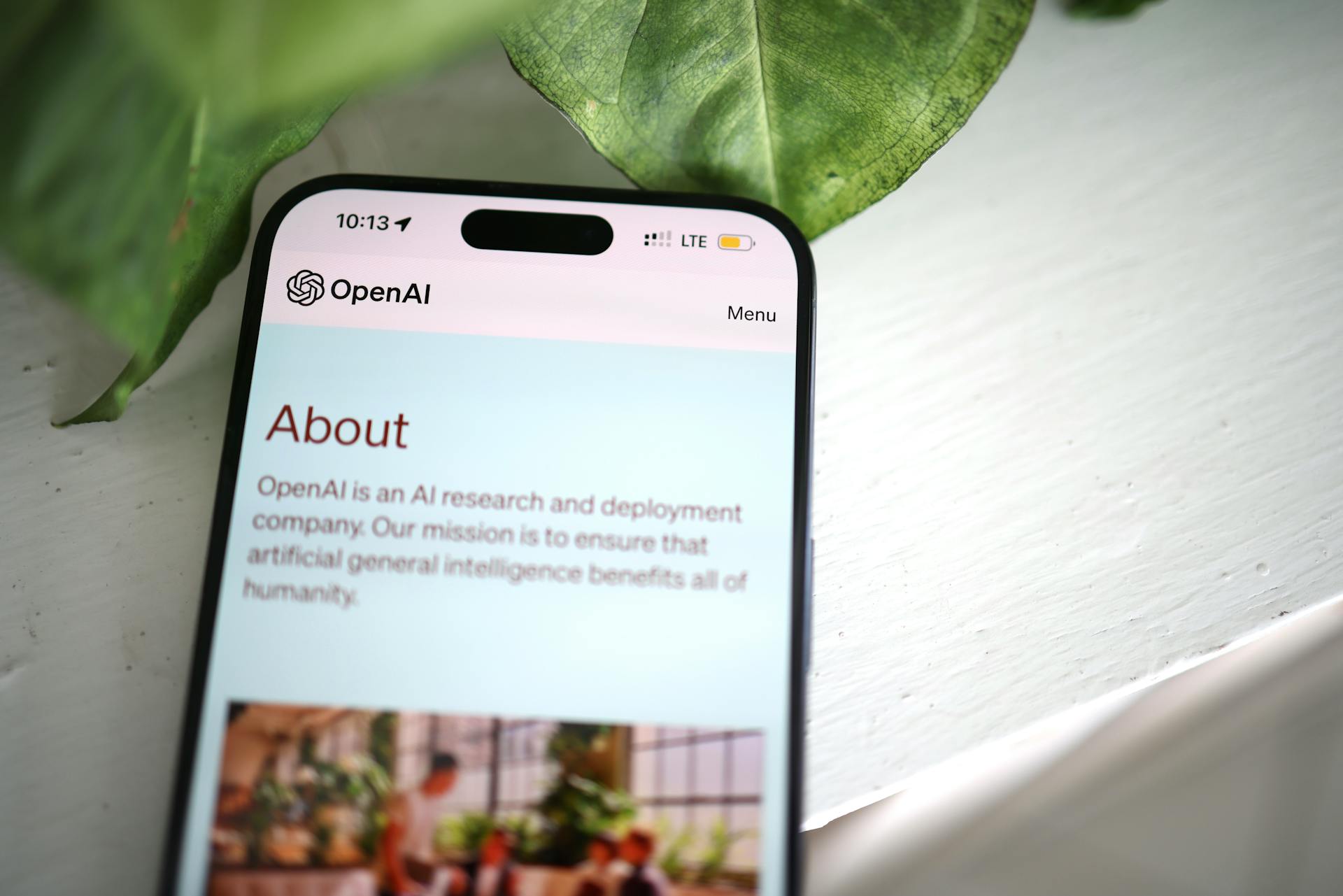 Beyaz bir masanın üzerinde duran telefonun ekranında OpenAI'ın websitesindeki Hakkında sayfası görülüyor. Telefonun arkasında bir bitkinin yeşil yaprakları var.