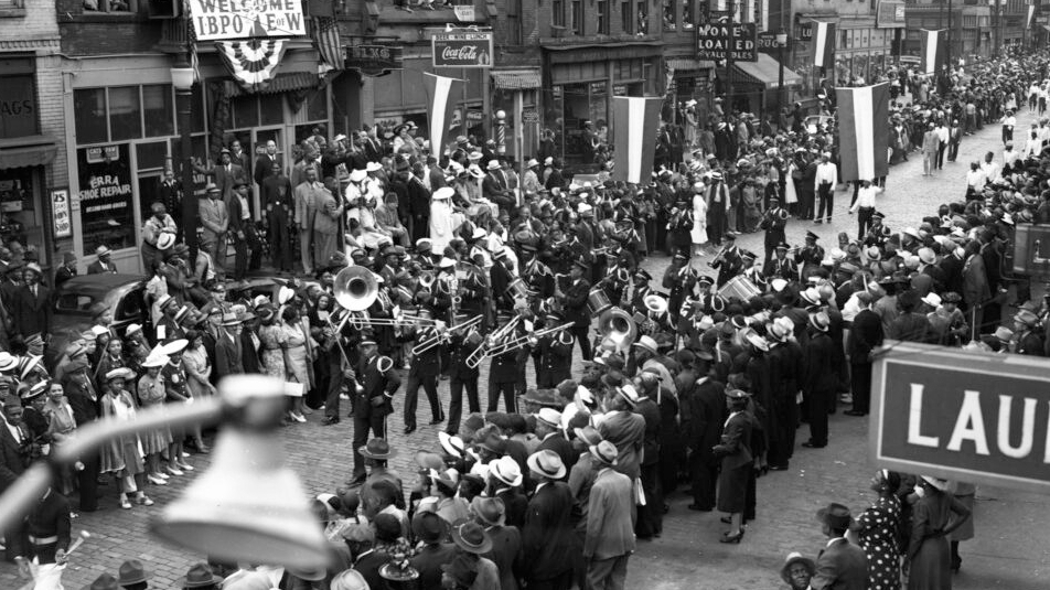 Black and white photo of people on the street. A group of people in the middle holding instruments as spectators watch.
